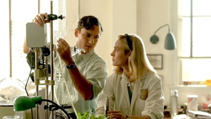 Image of Lewis Pullman as Calvin and Brie Larson as Elizabeth in a scene from 'Lessons in Chemistry' on Apple TV+. Calvin is a white man with short brown hair wearing a white lab coat. Elizabeth is a white woman with shoulder-length blonde hair wearing a white lab coat. They are both standing at a counter in a chemistry lab looking at each other as Calvin adjusts a fluid bag on a stand.